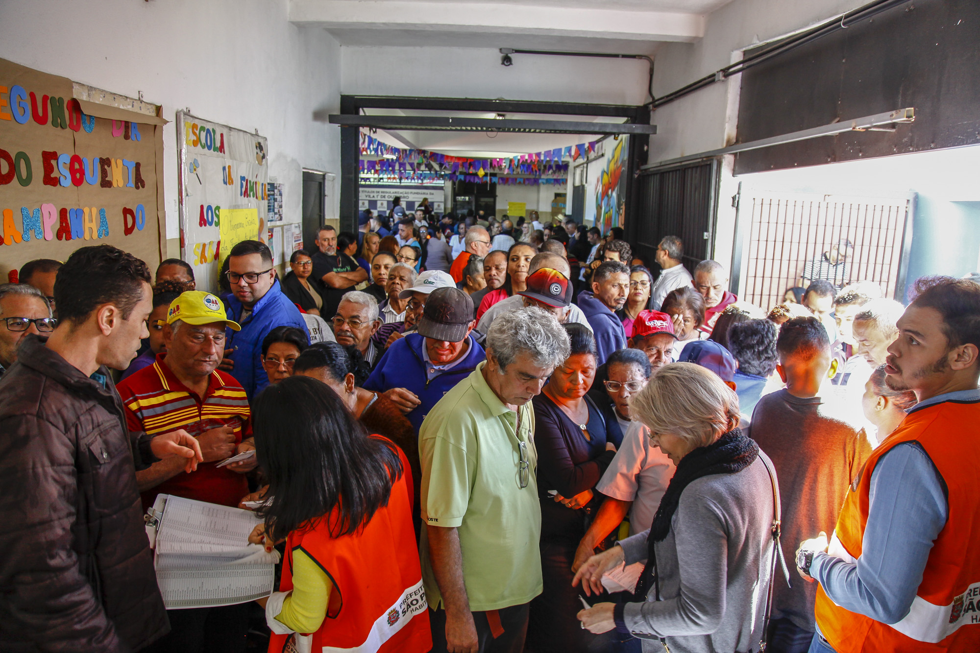 Foto tirada de cima, do corredor da escola que mostra os mutuários esperando a entrega dos mais de 900 títulos de propriedades. 