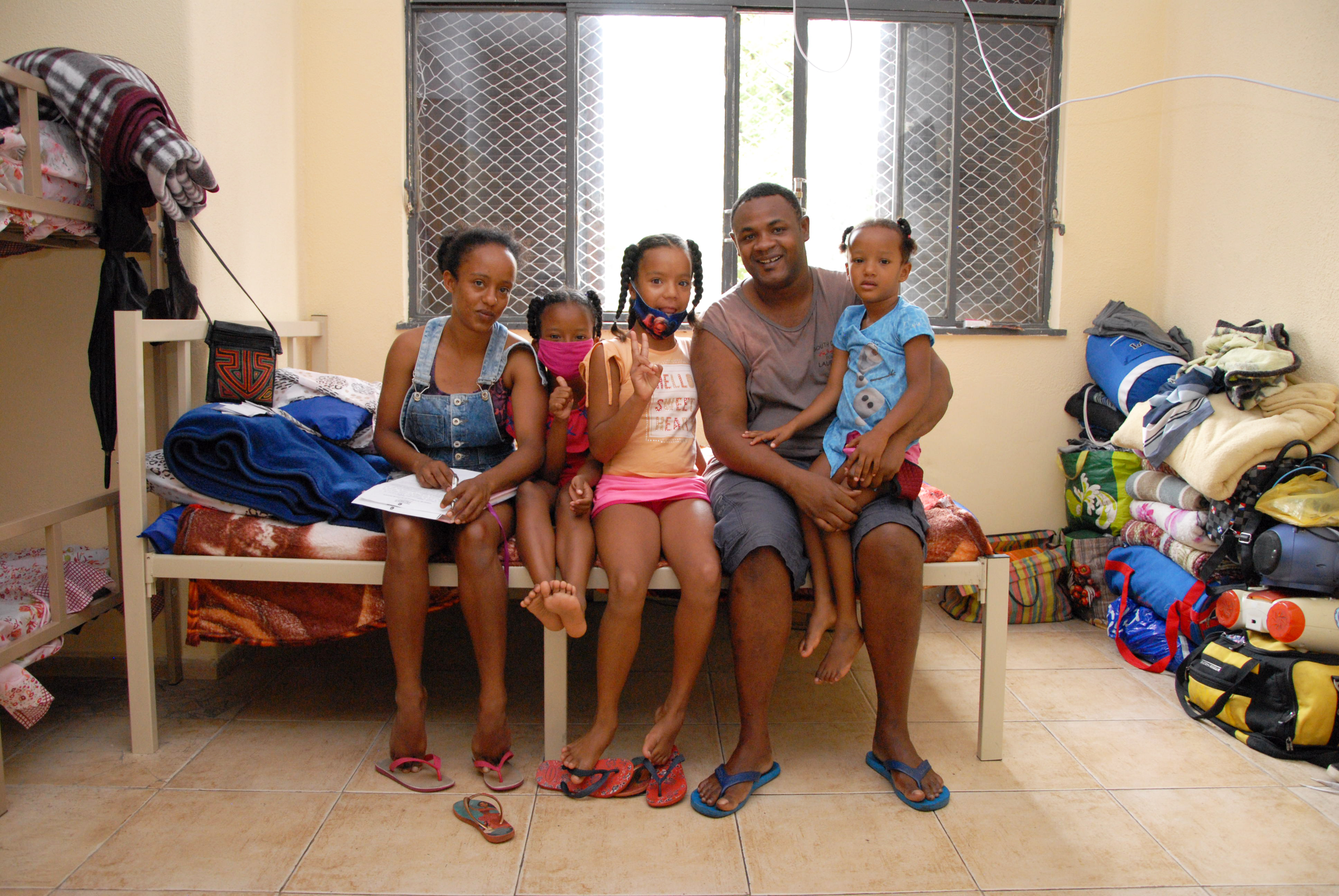 Família sentada na cama de sua nova residência. A família é constituída por mulher, marido e três filhas