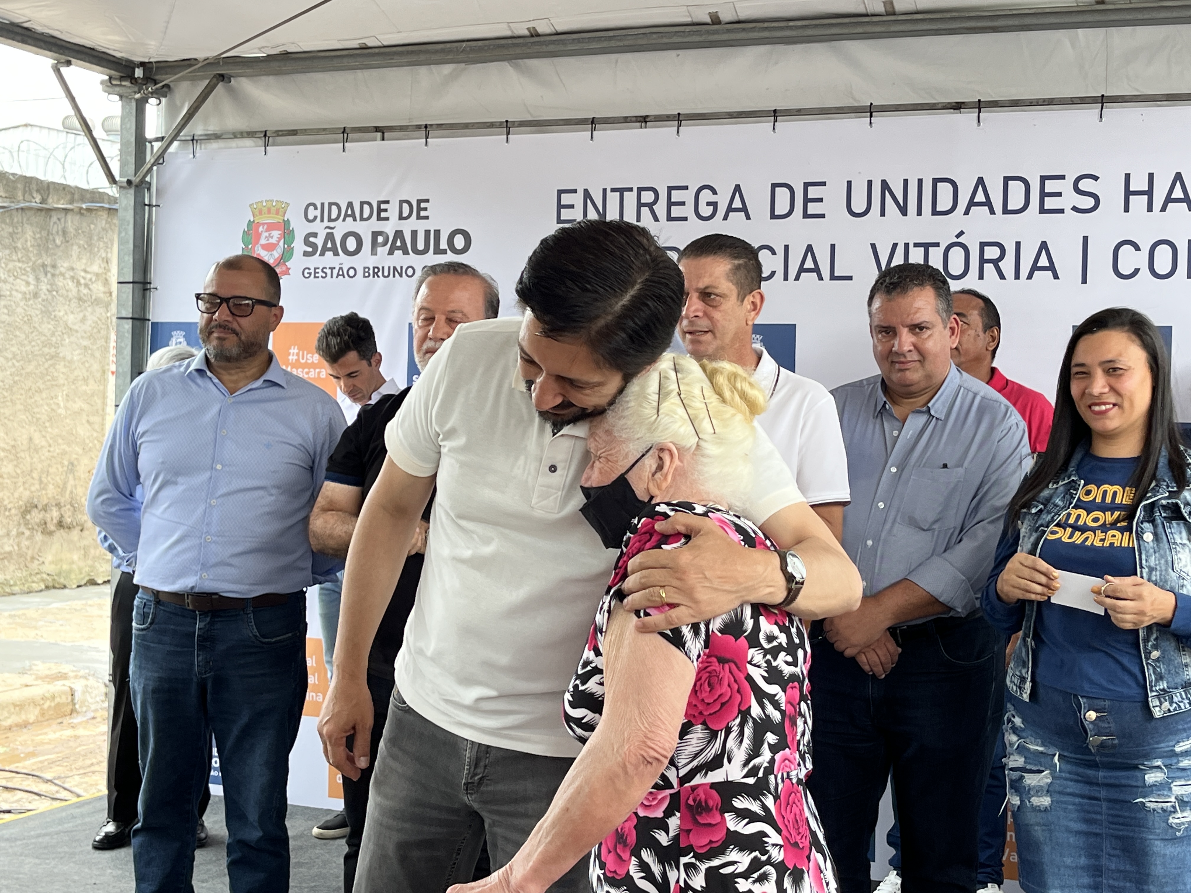 FOTO 02: Em primeiro plano, Prefeito Ricardo Nunes abraça senhora, em cima do palco do evento, em segundo plano o Secretário Municipal de Habitação João Farias