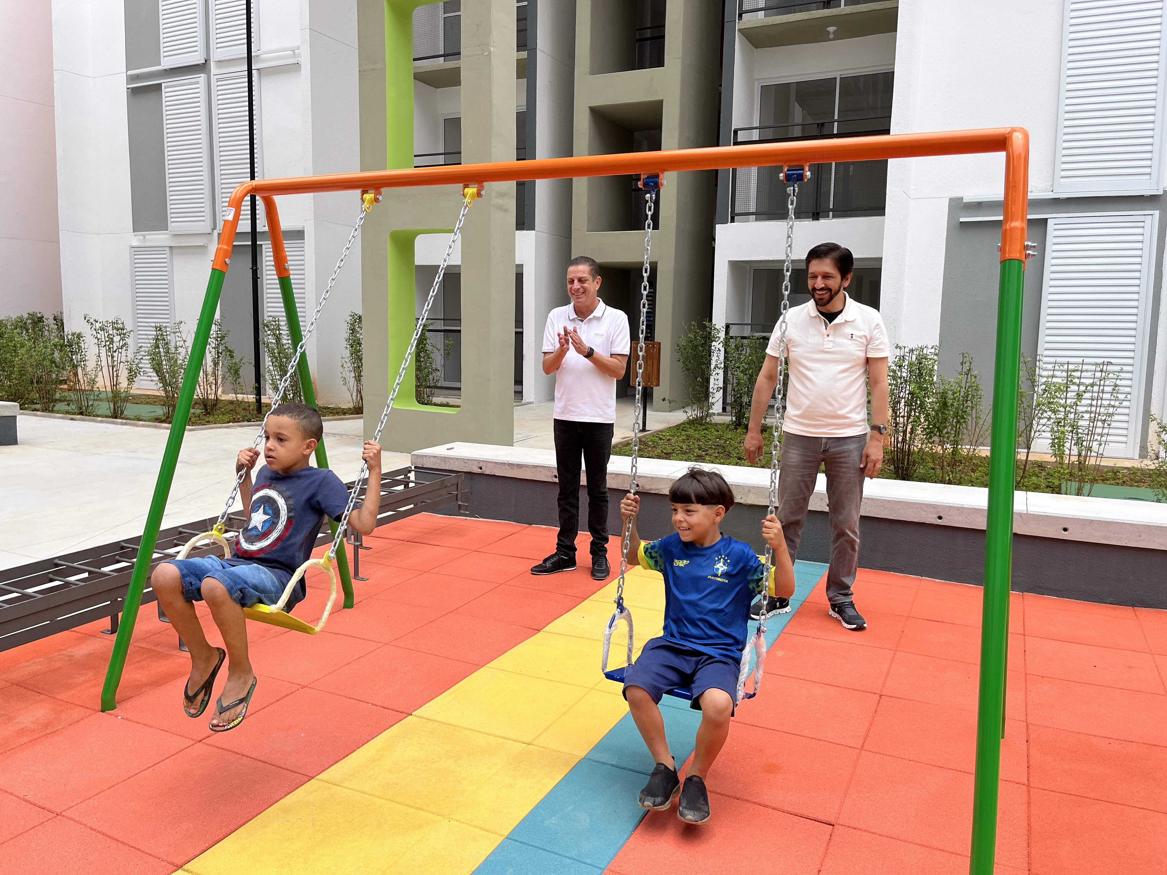 FOTO 01: Secretário Municipal de Habitação João Farias e Prefeito Ricardo Nunes brincando com duas crianças no balanço na área de playground