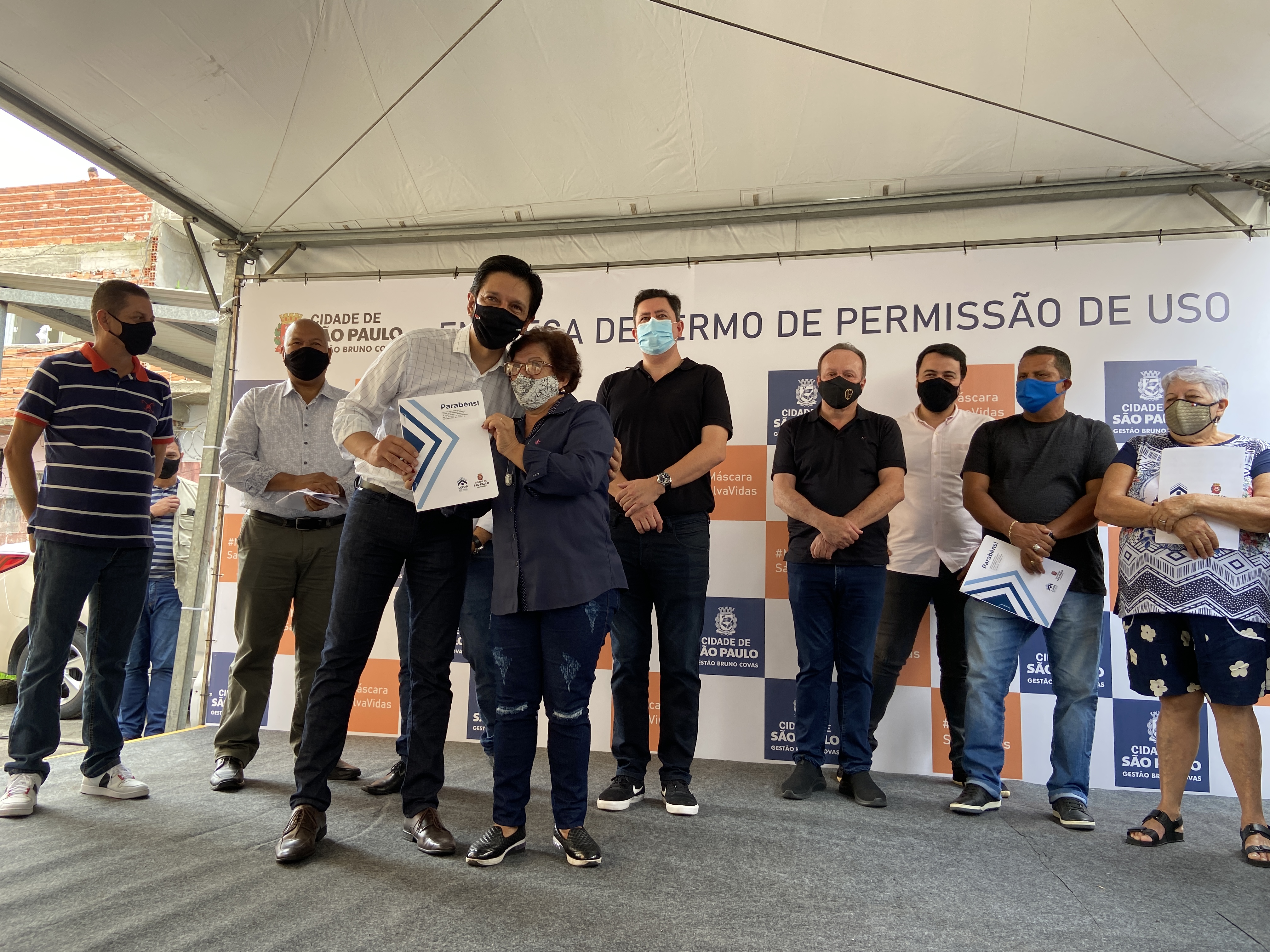 No palco estão algumas autoridades e o Prefeito Ricardo Nunes está entregando o Instrumento Contratual para uma das moradoras do Conjunto.