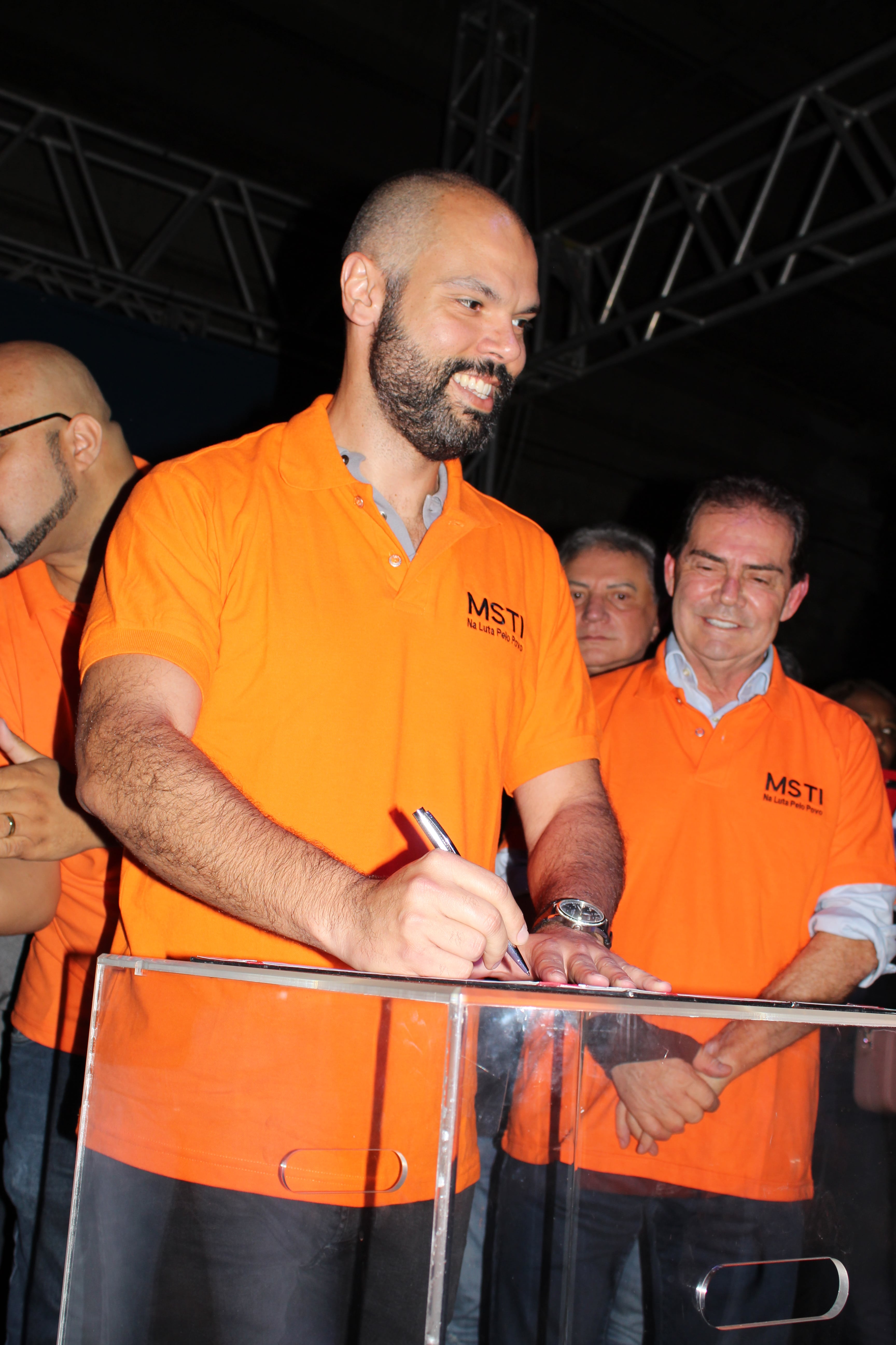 Na imagem, mostra o prefeito Bruno Covas vestido com a camiseta do MSTI, sorrindo e assinando um documento. 