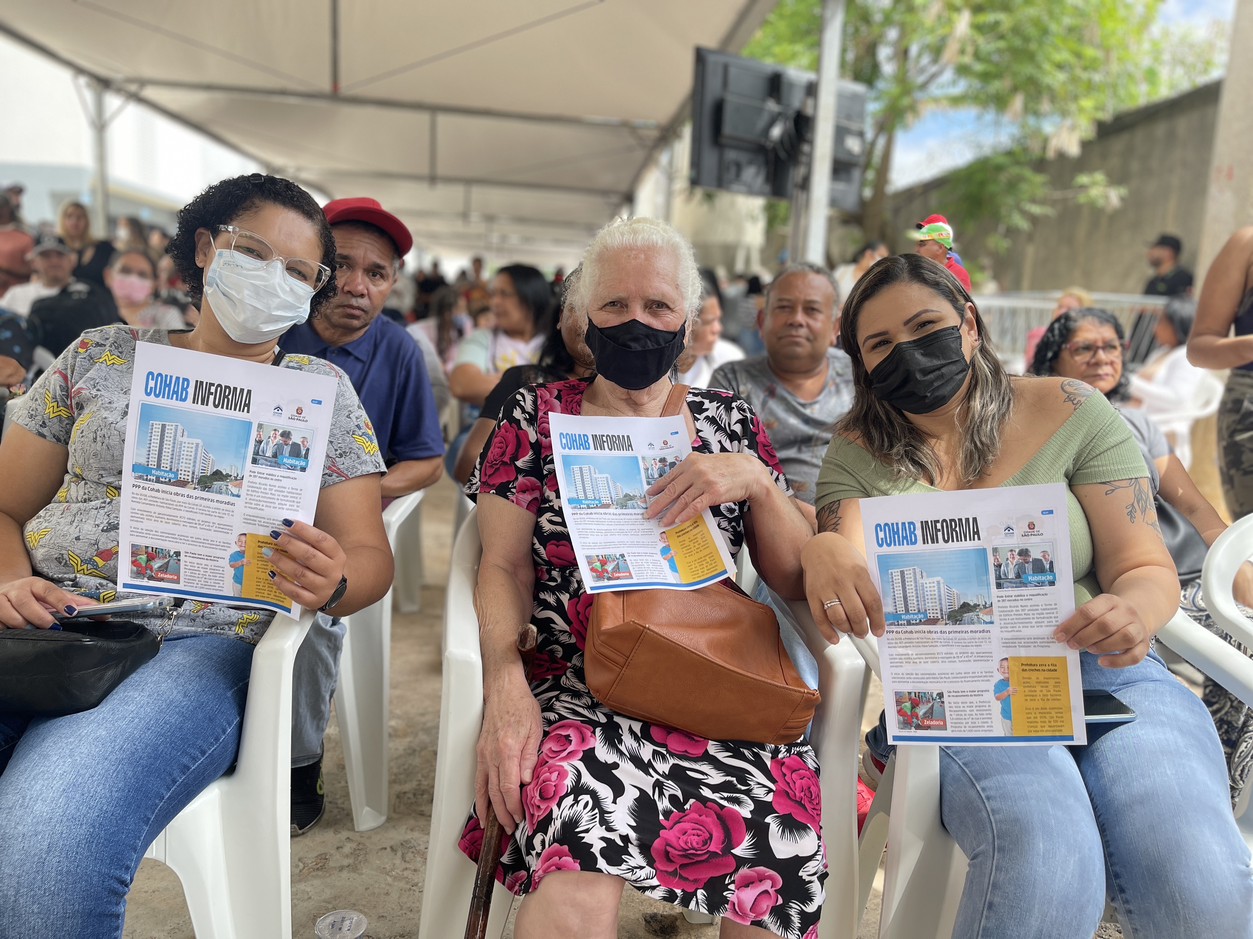 CAPA: Três senhoras sentadas segurando o jornal “Cohab Informa”  no evento de entrega do Complexo Habitacional em Heliópolis 