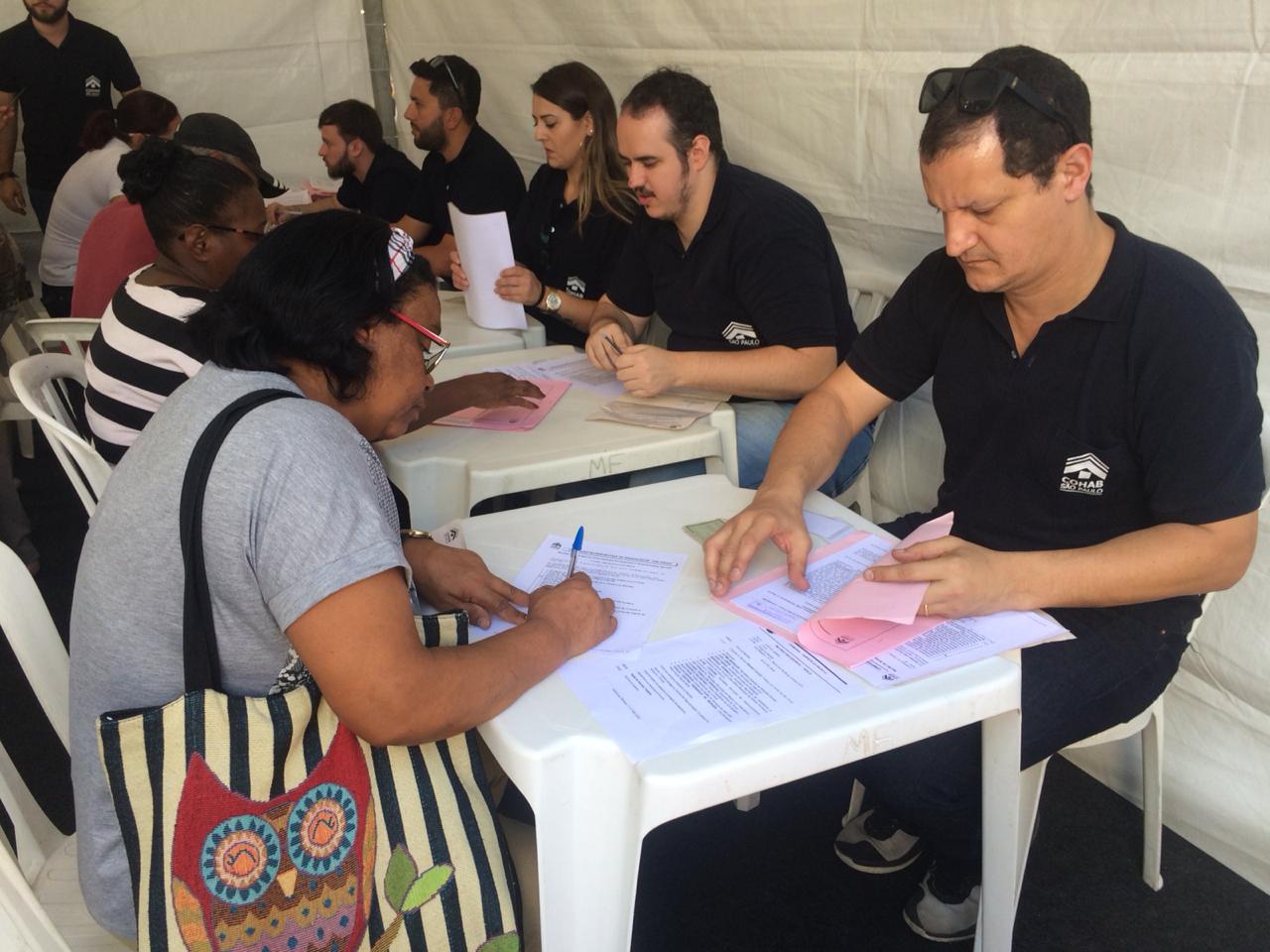 A imagem foi tirada em diagonal, mostrando toda a mesa dentro da tenda da Cohab. Com representantes sentados de frente para mutuários tirando dúvidas e assinando contratos. 