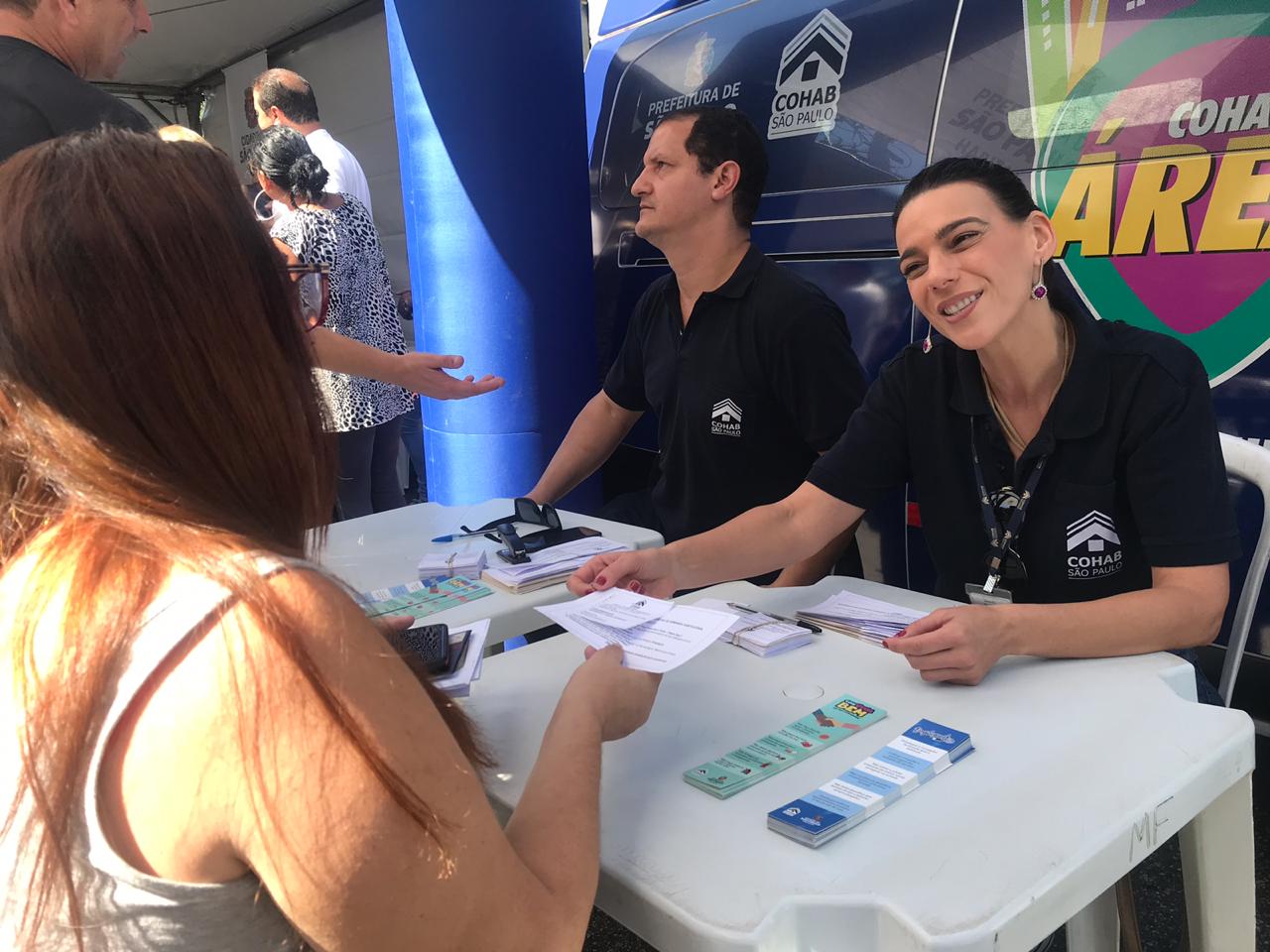 Na foto, ao fundo está a van "Cohab na Área" e sentados estão dois representantes da Cohab atendendo mutuários. 