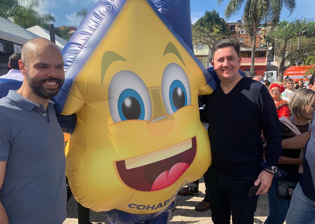 Ao lado esquerdo da foto está o prefeito de São Paulo Bruno Covas, ao meio está o mascote Cohabinho e ao lado está o presidente da Cohab Alexsandro Peixe. 