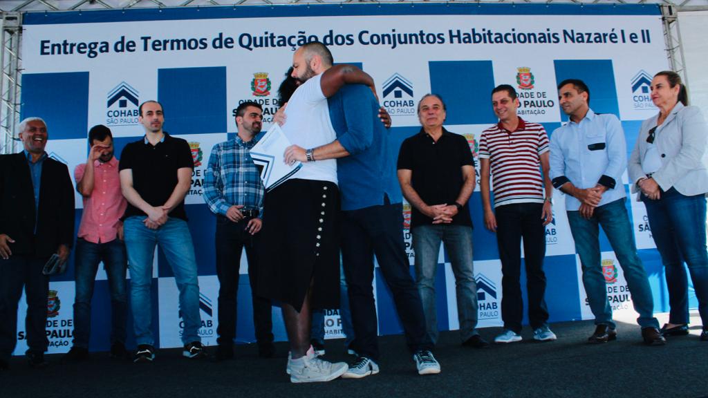 Na imagem está uma mutuária pegando seu termo de quitação em cima do palco e abraçando o prefeito de São Paulo Bruno Covas. 