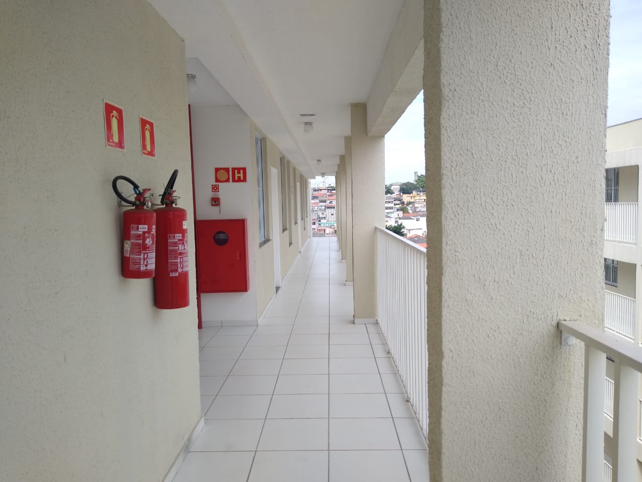 Foto do corredor do empreendimento. No primeiro plano estão extintores de incêndio. As paredes possuem a cor creme, com o chão sendo branco