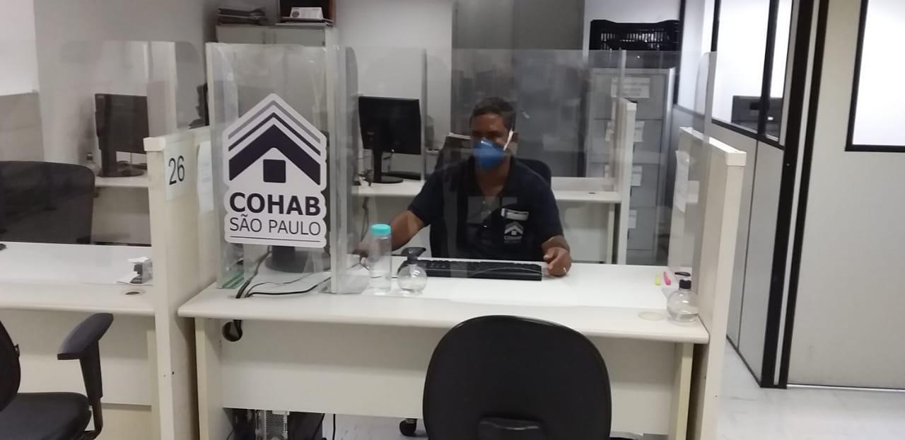 Foto com funcionário da Cohab sentado atrás de um vidro de isolamento com o logo da Cohab-SP. O funcionário está fazendo uso de um computador e está com uma máscara de proteção.