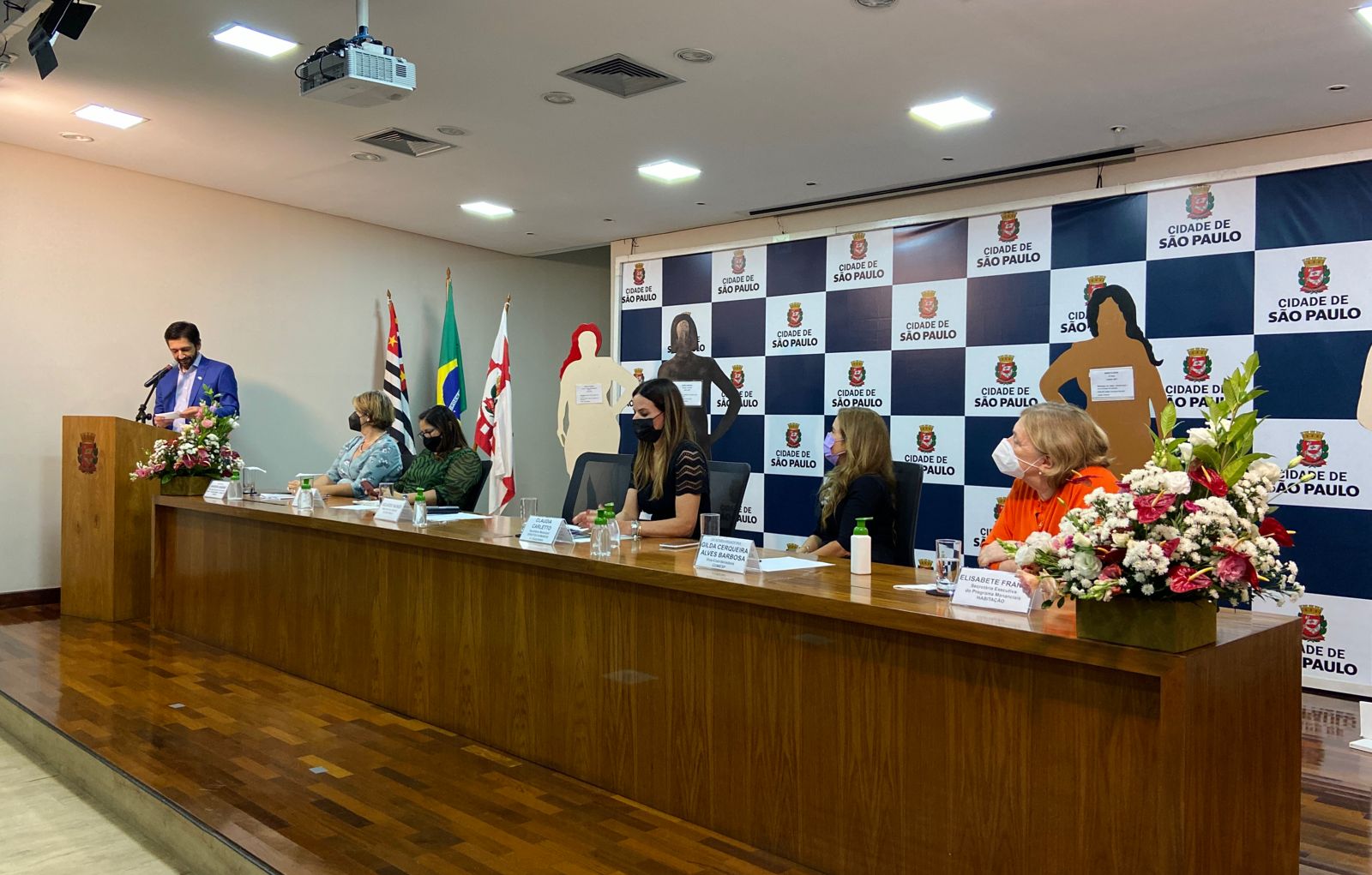 Prefeito Ricardo Nunes falando no púlpito, em seguida as secretárias mulheres estão sentadas compondo a mesa. 
