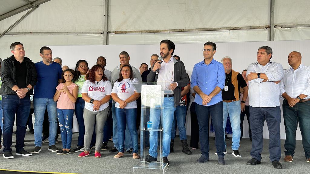 Prefeito Ricardo Nunes discursa no palanque enquanto outras autoridades e líderes comunitários assistem atrás dele
