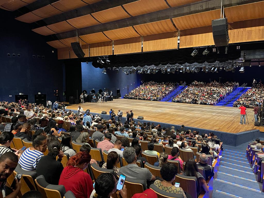 A imagem mostra um auditório com pessoas nas poltronas, virado a um palco