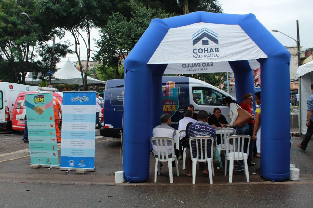 No lado esquerdo da imagem está os banners do Conviva Bem e Desplugando. Ao centro da foto está a tenda da Cohab com dois representantes da Companhia sentados com dois mutuários entregando os termos de quitação. Além de ter dois de costas esperando para serem atendidos. 